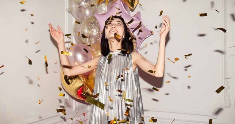 A woman in a silver dress joyfully throws gold confetti in the air. Behind her are star-shaped and round balloons in metallic colors, set against a white wall. She appears to be celebrating, with a wide smile and arms raised.