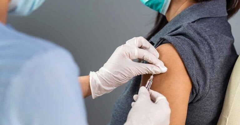 A healthcare professional administers a vaccine to a person's arm. Both individuals are wearing face masks, and the person receiving the vaccine is seated, wearing a dark gray shirt.