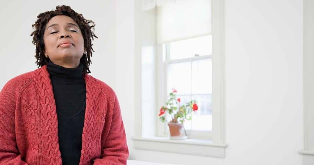A person in a red sweater and black turtleneck sits calmly with their eyes closed. In the background, a window lets in natural light, with a potted plant on the sill. The room has white walls, creating a peaceful ambiance.