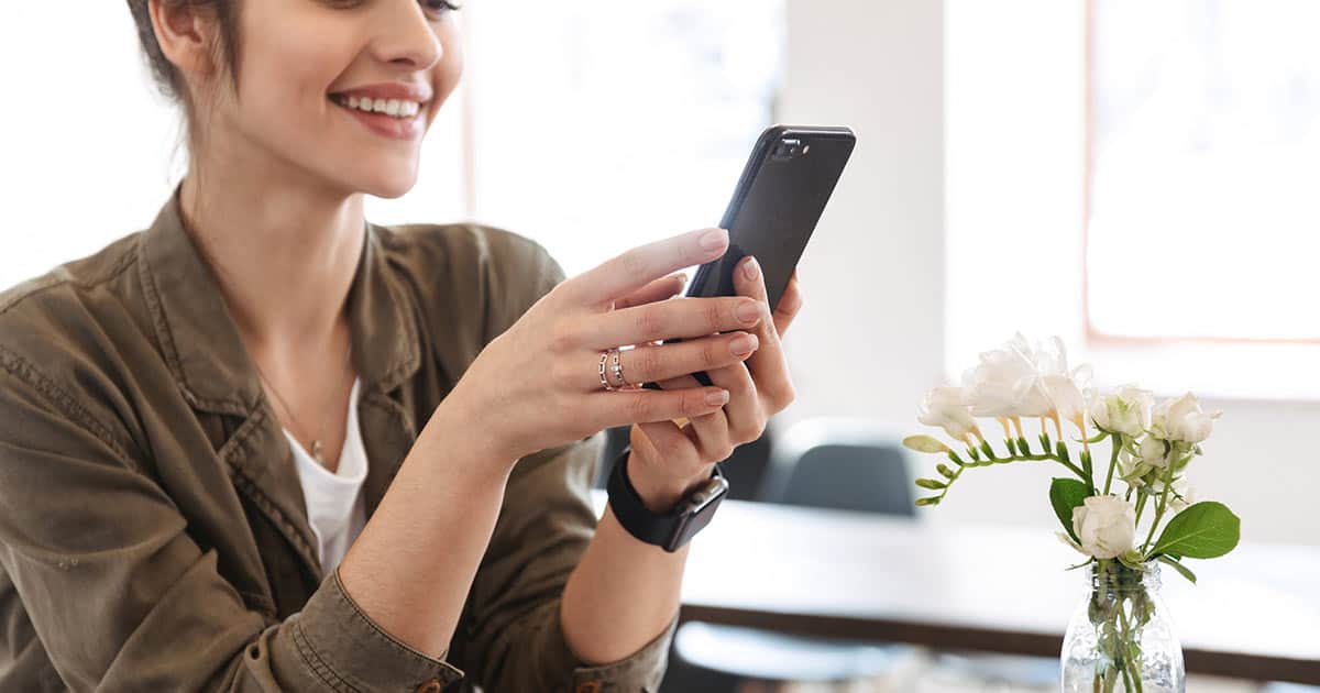 A person smiling while looking at a smartphone, sitting at a table with a small vase of white flowers. The background is softly lit, suggesting a casual and pleasant indoor setting.