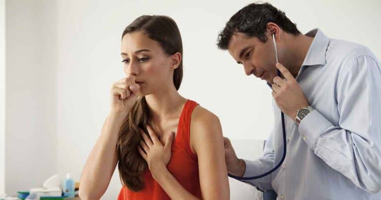 A woman in a red tank top is coughing, covering her mouth with one hand and holding her chest with the other. A man in a light blue shirt, wearing a stethoscope, is listening to her back, indicating a medical examination.