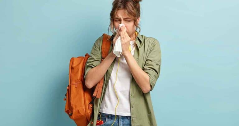 A person with a backpack stands against a light blue background, sneezing or blowing their nose into a tissue. They are wearing a green jacket over a white t-shirt and have an expression of discomfort.