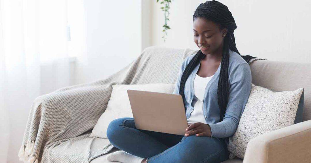 A woman with long braided hair sits cross-legged on a couch, using a laptop. She wears a blue cardigan and jeans, smiling as she looks at the screen. Natural light brightens the room, and a cozy blanket drapes over the sofa.