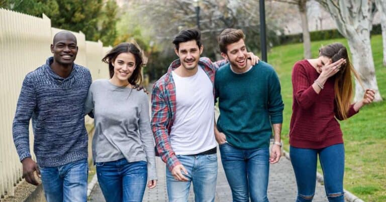 A group of five friends walking outdoors, smiling and casually dressed. Two men and two women have their arms around each other, while one woman covers her face playfully. They are on a paved path with grassy areas and trees in the background.