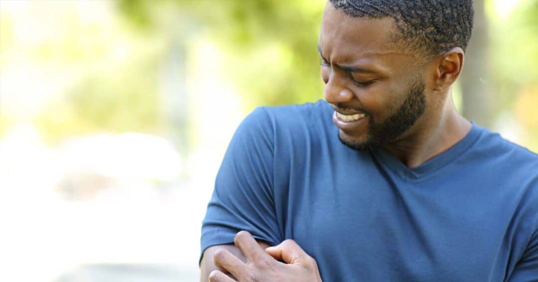 A person in a blue shirt is outdoors, grimacing and touching their upper arm with their right hand, as if experiencing discomfort. The background is blurred, suggesting a sunny day with greenery.