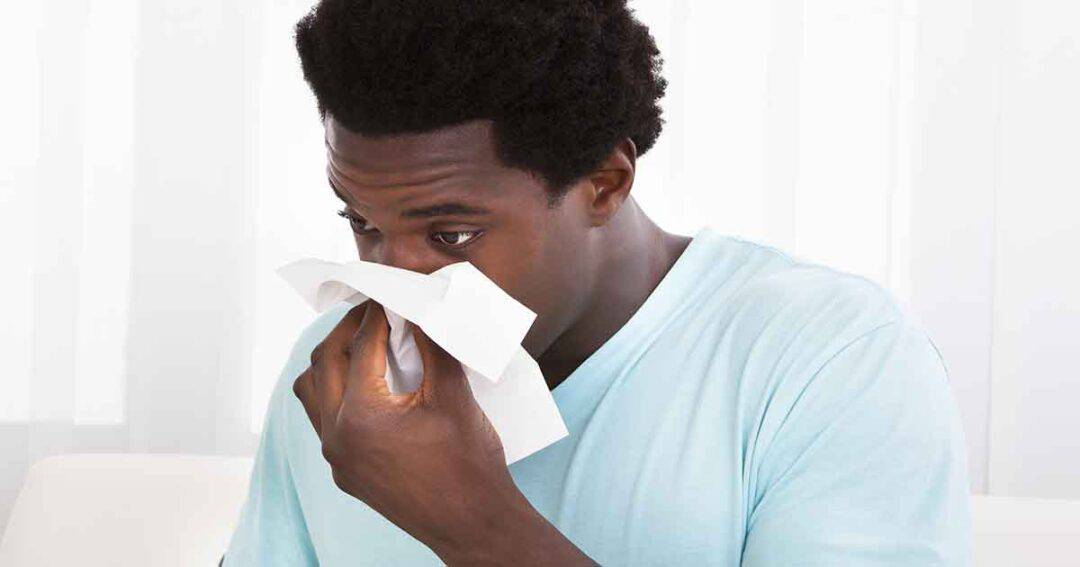 A man wearing a light blue shirt sits indoors, holding a tissue to his nose as if sneezing or wiping it. The background is softly lit with white curtains.