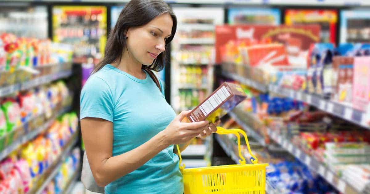 A woman in a blue shirt is shopping in a grocery store aisle, holding a yellow basket. She is reading the label on a boxed product while surrounded by colorful packaged goods on the shelves.