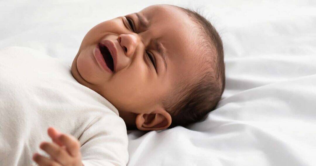 A baby lying on a white bed, wearing a light-colored outfit, with eyes closed and mouth open, appears to be crying or upset, which could be an early sign of infant anaphylaxis.