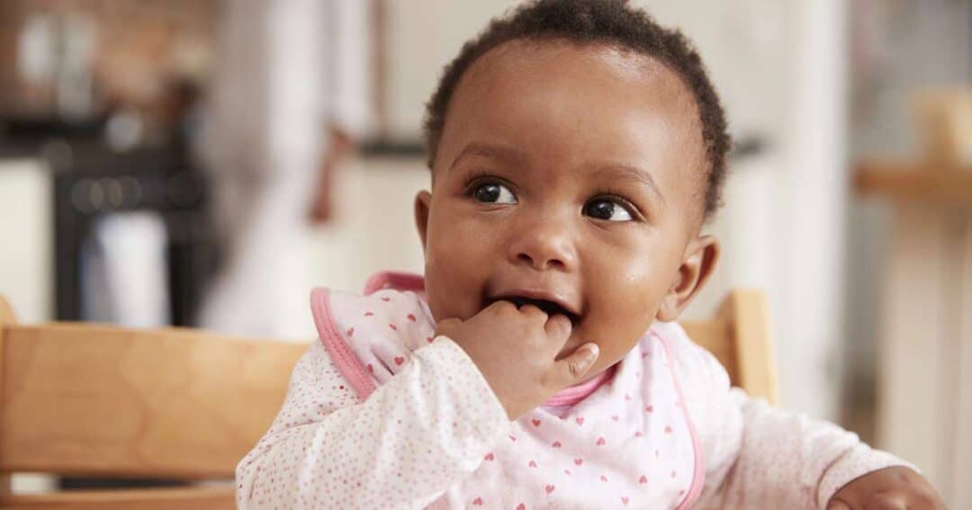 A baby wearing a pink and white outfit sits in a high chair, smiling and chewing on their fingers—an adorable moment that reminds us to watch for signs of infant anaphylaxis. The softly blurred background keeps the focus on the baby.
