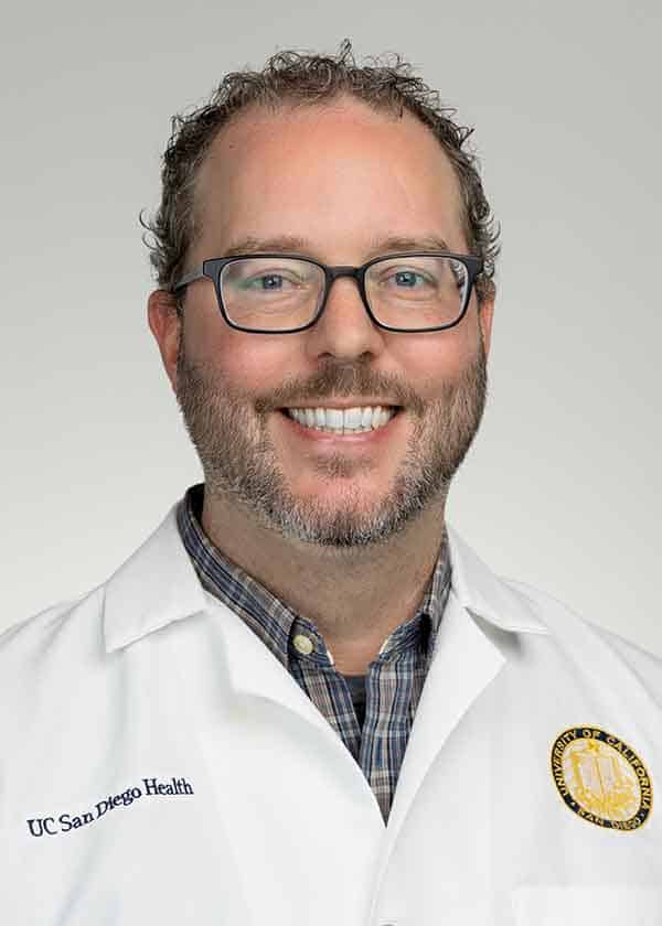 A smiling man with short curly hair, glasses, and a beard wears a white lab coat with the UC San Diego Health logo and a plaid shirt, standing against a light gray background.