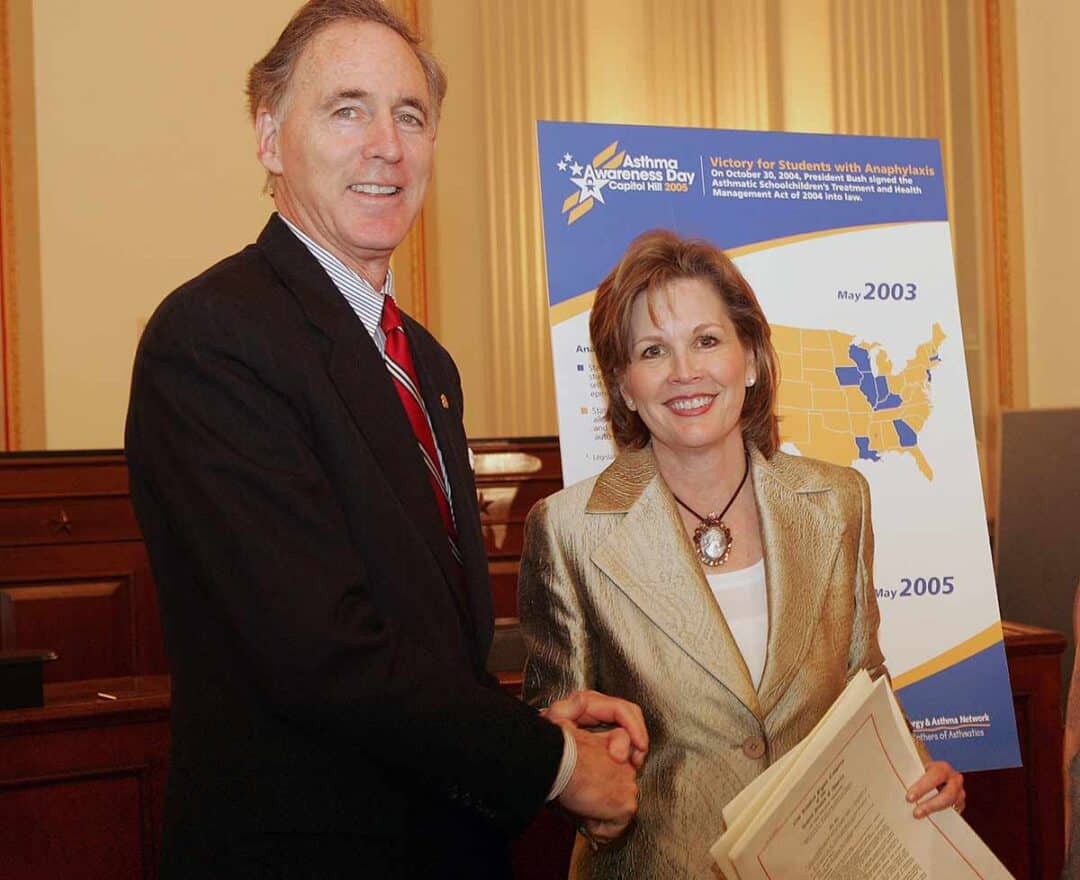 A man in a suit and a woman in a gold blazer shake hands and smile at an indoor event. Behind them is a poster about Asthma Awareness Day and student anaphylaxis laws with a U.S. map and timeline.