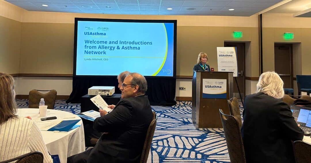 A woman stands at a podium giving a presentation titled “Welcome and Introductions from Allergy & Asthma Network” to an audience seated in a conference room. Attendees listen and look at papers.