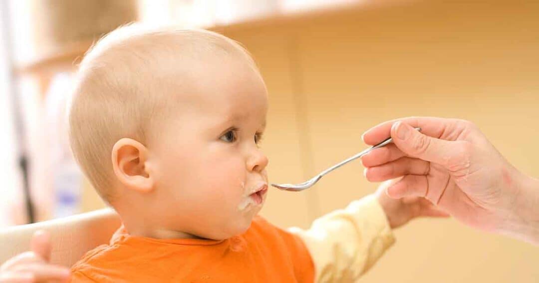 A baby in an orange bib sits in a high chair while an adult hand holds a spoon, feeding the baby. The baby looks to the side with a bit of food on their lips. The background is softly blurred.
