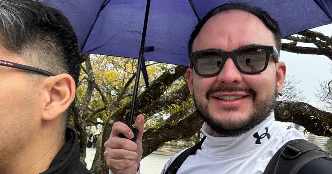 Two people are outside; one is smiling at the camera under a blue umbrella, wearing sunglasses and a white jacket. The other person is partly out of frame. Leafy trees and a cloudy sky are in the background.
