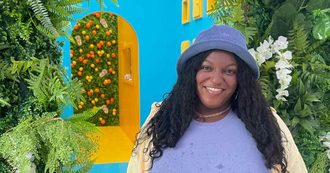 A woman with long curly hair and a blue bucket hat smiles in front of a colorful backdrop with green plants and a bright blue and yellow wall decorated with citrus fruits and flowers.