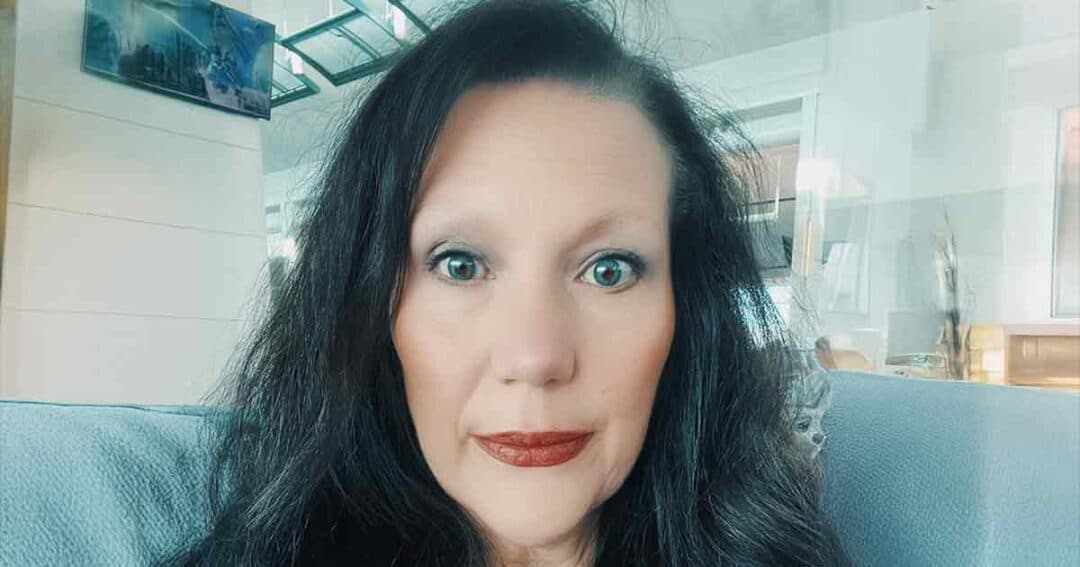 A woman with long dark hair and light eyes sits indoors on a light blue couch, looking directly at the camera with a neutral expression. The background shows modern home decor and glass reflections.