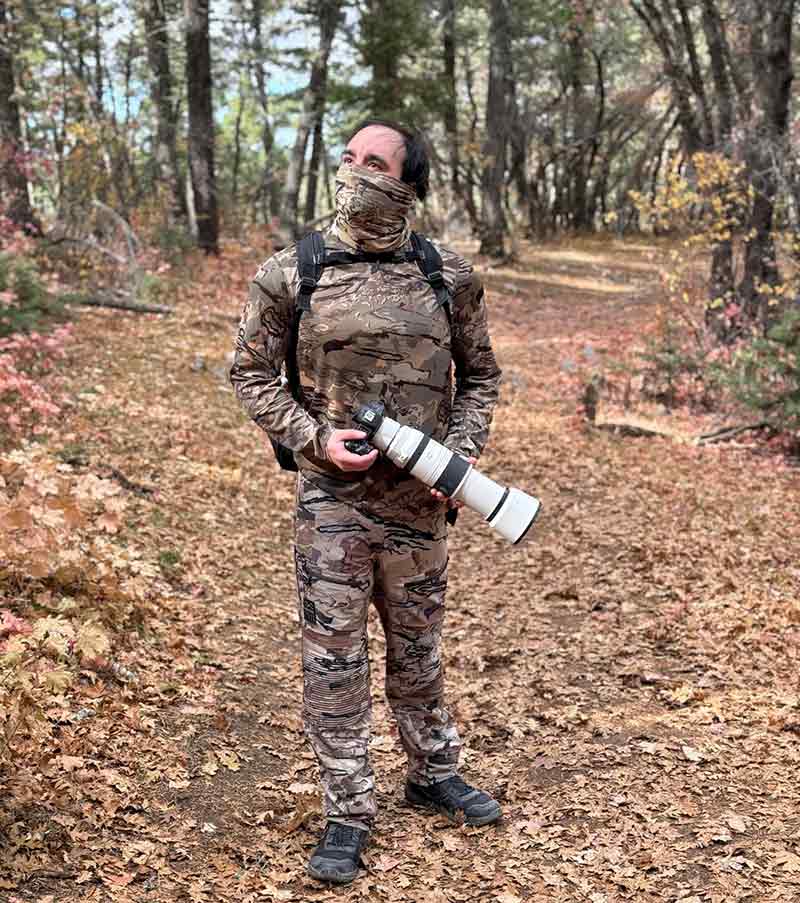 A person in full camouflage clothing stands on a forest trail holding a large camera with a telephoto lens. Autumn leaves cover the ground and trees with colorful foliage surround the scene.