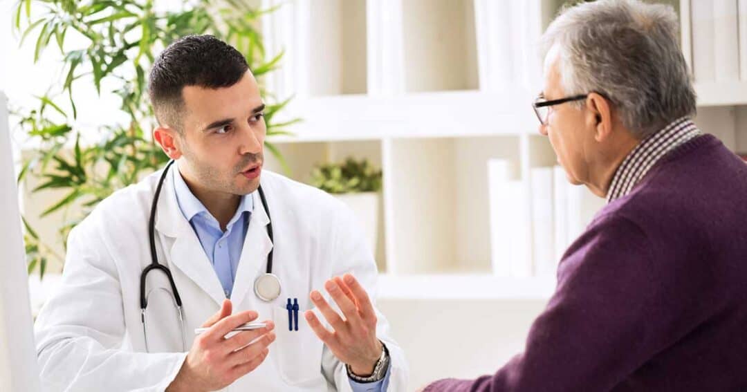 A doctor in a white coat with a stethoscope talks to an older man in glasses and a purple sweater during a medical consultation in a bright office.