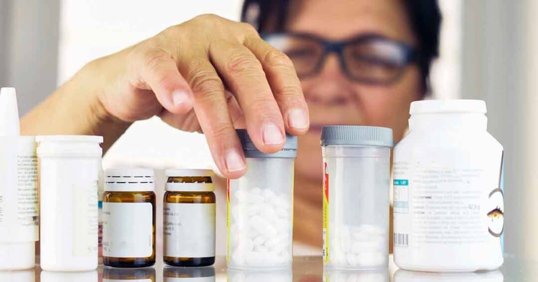 A person wearing glasses reaches for a pill bottle among several different medication containers arranged in a row on a surface.