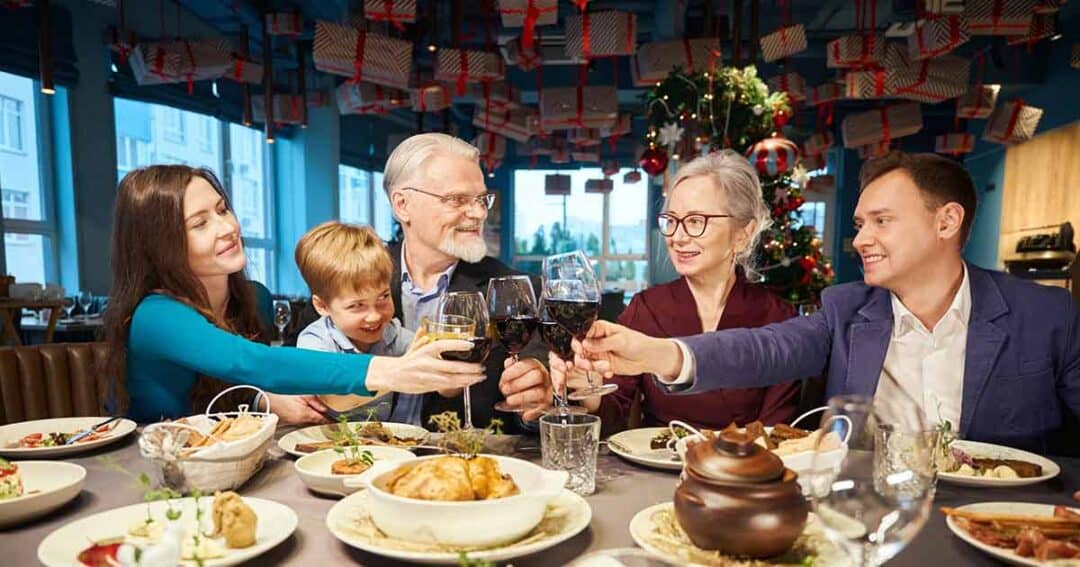 A family of five sits around a festive dinner table, raising glasses in a toast. There is food on the table, and a decorated Christmas tree with gifts in the background. Everyone is smiling and enjoying the celebration.