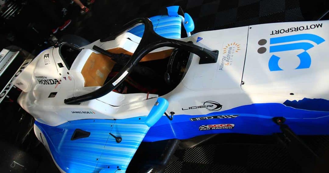 A top view of a blue and white race car with visible sponsor logos, a Honda emblem, and a transparent driver's seat cover, parked on a checkered floor.