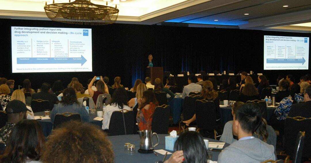 A speaker presents a slide about patient input in drug development to a seated audience in a large conference room with chandeliers. Attendees face the stage, listening and taking notes.