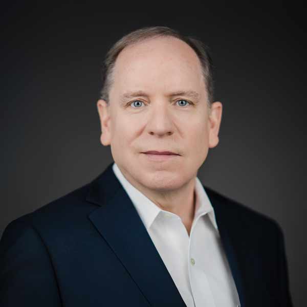 A middle-aged man with light skin, short light brown hair, and blue eyes wears a dark suit jacket and white collared shirt, posing against a plain dark background.