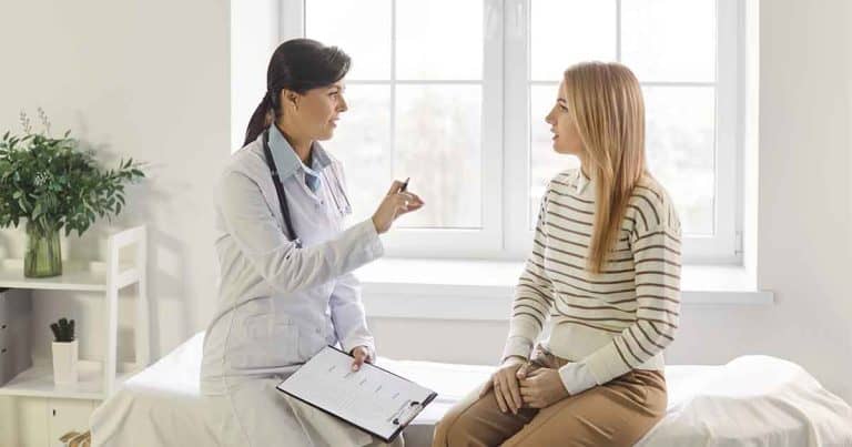 A doctor holding a clipboard talks to a woman sitting on an exam table in a bright medical office with a window in the background.