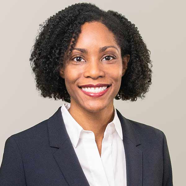 A woman with short curly hair is smiling and wearing a dark blazer over a white shirt, standing against a plain light background.