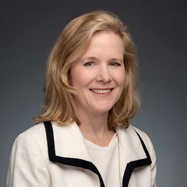 A woman with shoulder-length blonde hair, wearing a white blazer with black trim, smiles at the camera against a gray background.