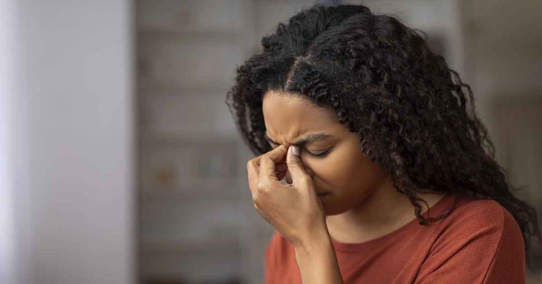A woman with curly hair wearing an orange top pinches the bridge of her nose and closes her eyes, appearing stressed or fatigued, indoors with a blurred background.
