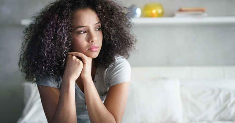 A young woman with curly hair sits on a bed, resting her chin on her hands and looking thoughtfully into the distance. The background is softly lit, with a shelf and pillows visible.