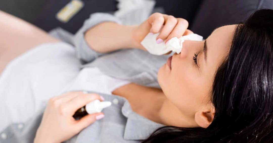 A woman reclines on a couch, holding a tissue to her nose and a nasal spray bottle in her hand, suggesting she has a cold or allergies.