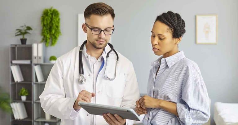 A doctor in a white coat with a stethoscope discusses information on a tablet with a patient in a bright medical office. The patient listens attentively. Shelves and plants are visible in the background.