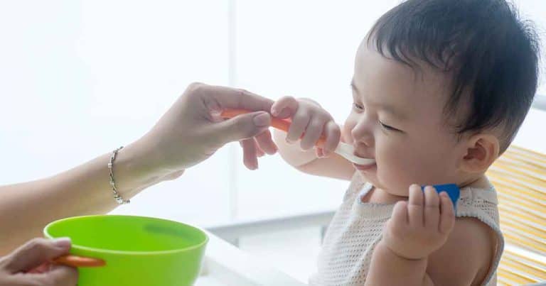 An adult feeds a baby with a spoon, while the baby holds another spoon and makes a face, possibly refusing the food. A green bowl is held in the adult’s other hand.