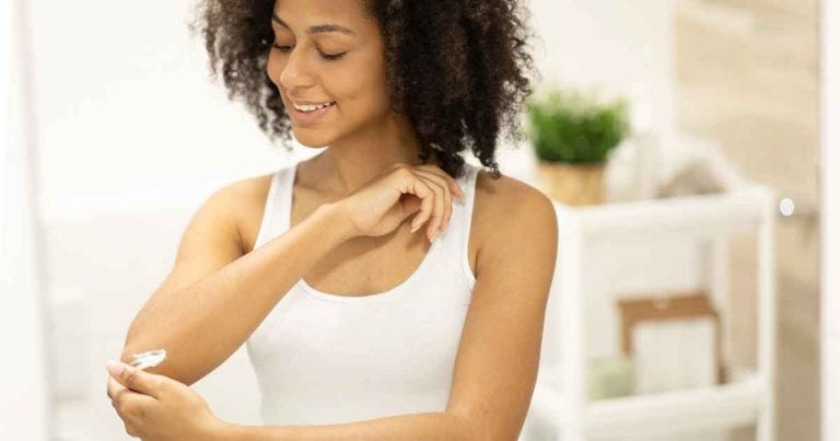 A woman with curly hair and a white tank top is smiling and applying lotion to her elbow in a bright bathroom.