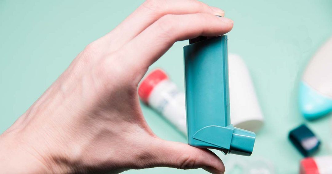 A hand holding a blue asthma inhaler against a light green background, with other medical items blurred in the background.
