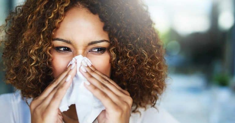 A woman with curly hair holds a tissue to her nose, appearing to sneeze or blow her nose, suggesting she might have a cold or allergies. The background is blurred.