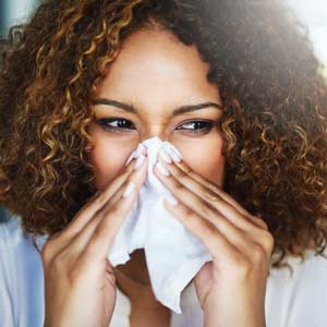 A person with curly hair holds a tissue to their nose, appearing to sneeze or wipe their nose, possibly due to a cold or allergies.