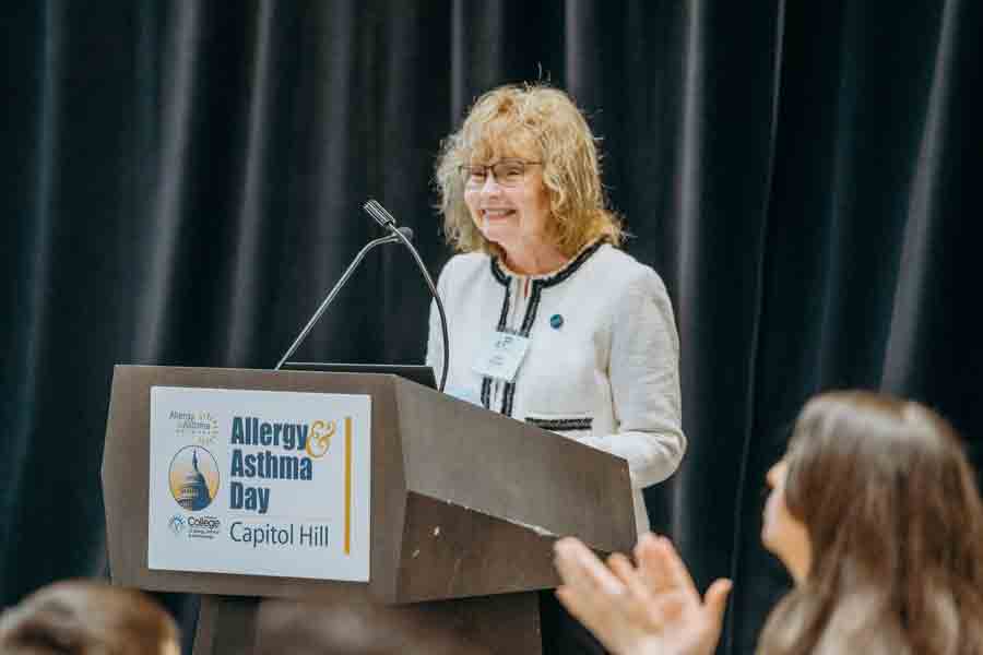 A woman with curly blonde hair and glasses speaks at a podium labeled "Allergy & Asthma Day, Capitol Hill," while audience members watch and applaud. A black curtain hangs in the background.