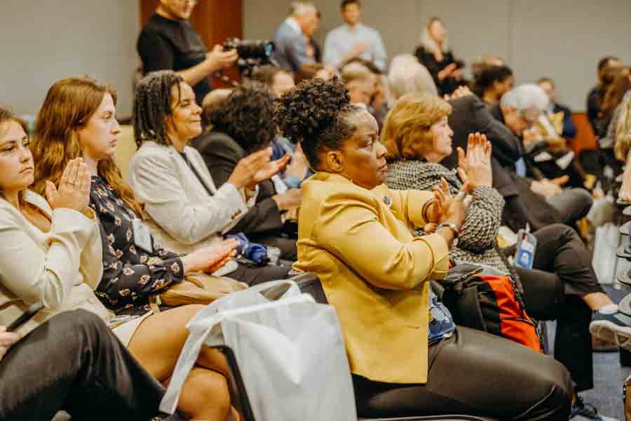 A diverse group of people sits closely together in a conference room, attentively listening and applauding. Some hold papers and bags, and the atmosphere is focused and engaged.