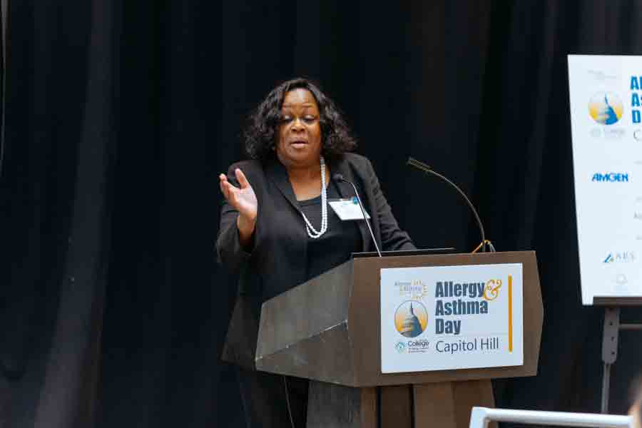 A woman stands at a podium speaking during an event called "Allergy & Asthma Day on Capitol Hill." She gestures with one hand, and a sign with event sponsors is visible beside her.