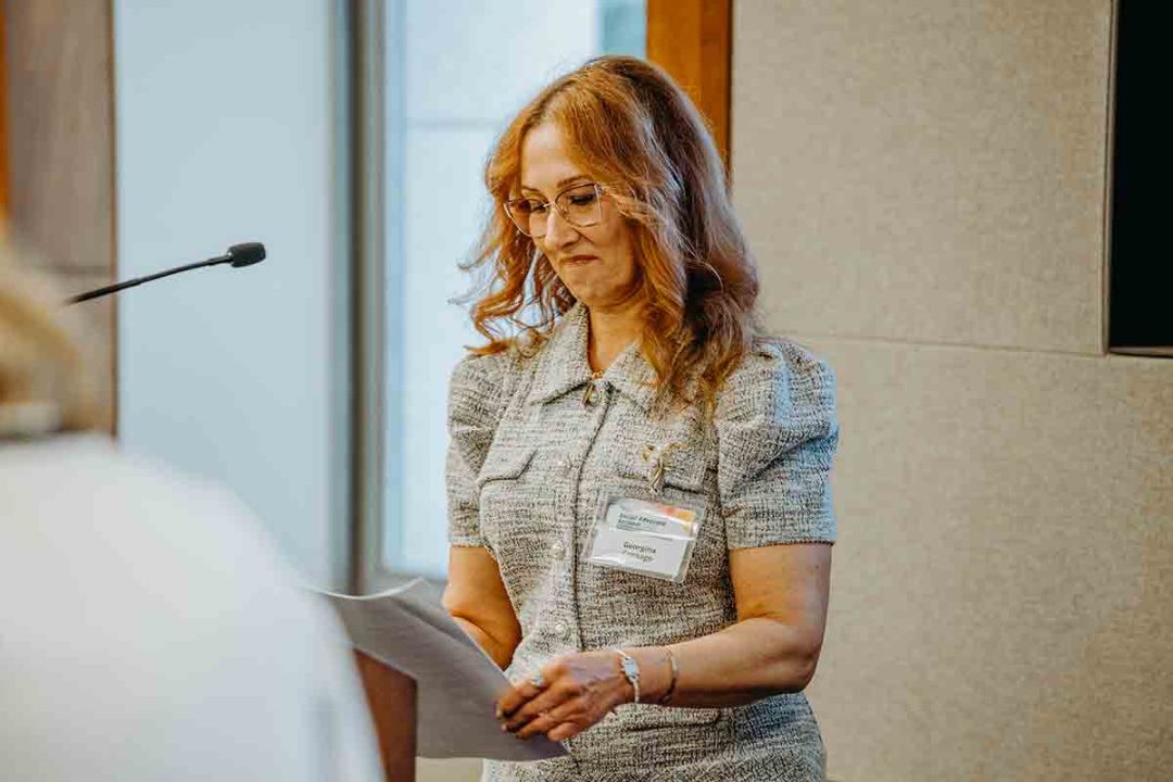 A woman with long, wavy hair and glasses stands indoors, reading from a sheet of paper. She wears a gray short-sleeved blouse and a name tag, and stands near a microphone.