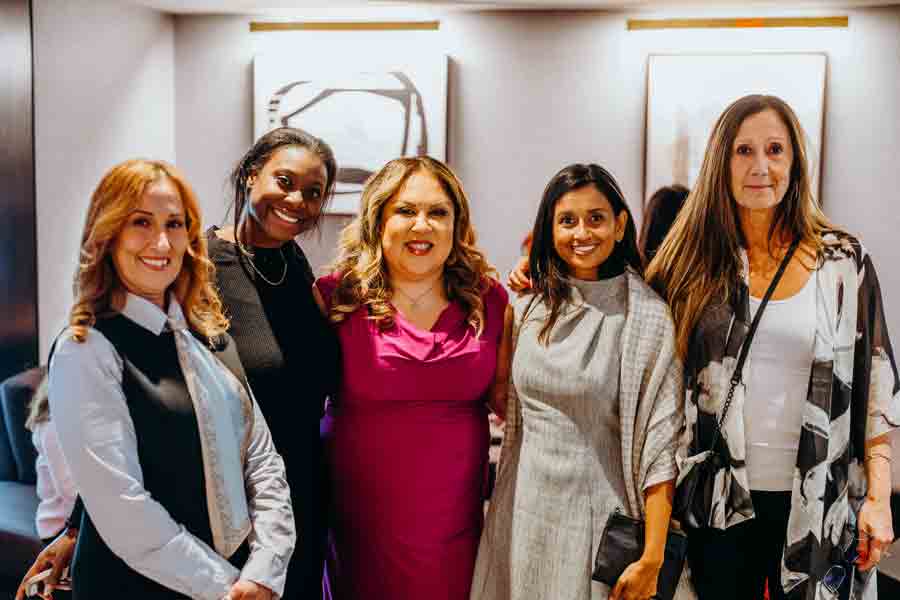 Five women stand together indoors, smiling at the camera. They are dressed in a mix of business and casual attire, with abstract artwork visible on the wall behind them.