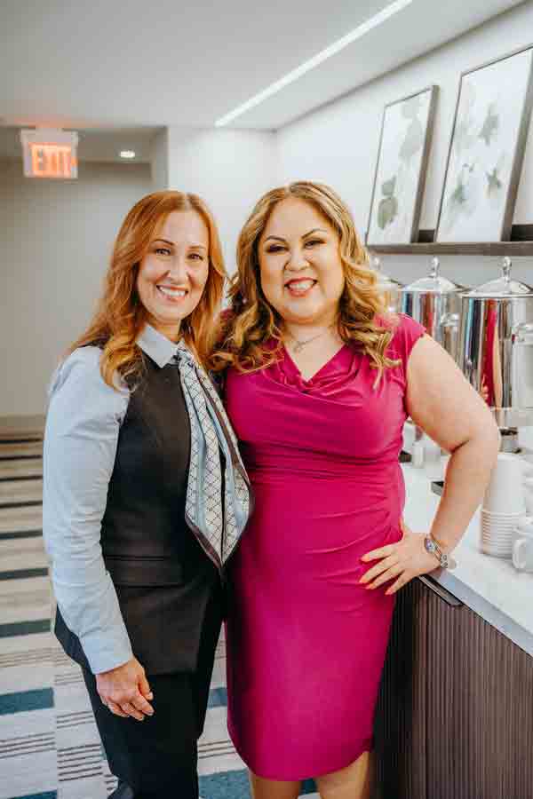 Two women smiling and posing together indoors; one wears a blue shirt with a vest and scarf, the other wears a bright pink dress. They stand by a counter with coffee dispensers and cups in a modern room.