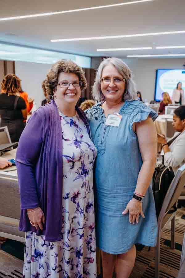 Two women smile and stand together in a conference room. One wears a purple floral dress and cardigan; the other wears a light blue dress and glasses. People are seated and a presentation screen is visible in the background.