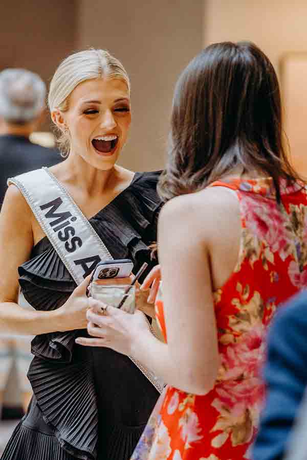 A woman wearing a "Miss" sash and a black dress smiles excitedly while talking to another woman in a red floral dress at an indoor event.