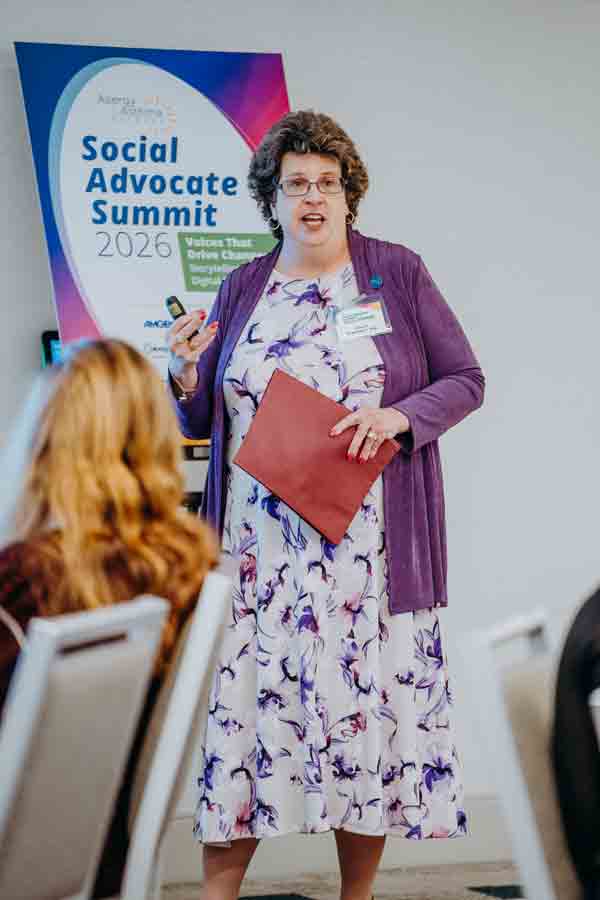 A woman in a floral dress and purple cardigan speaks at a podium during the Social Advocate Summit 2026, holding a red folder and microphone. A summit poster is displayed in the background.