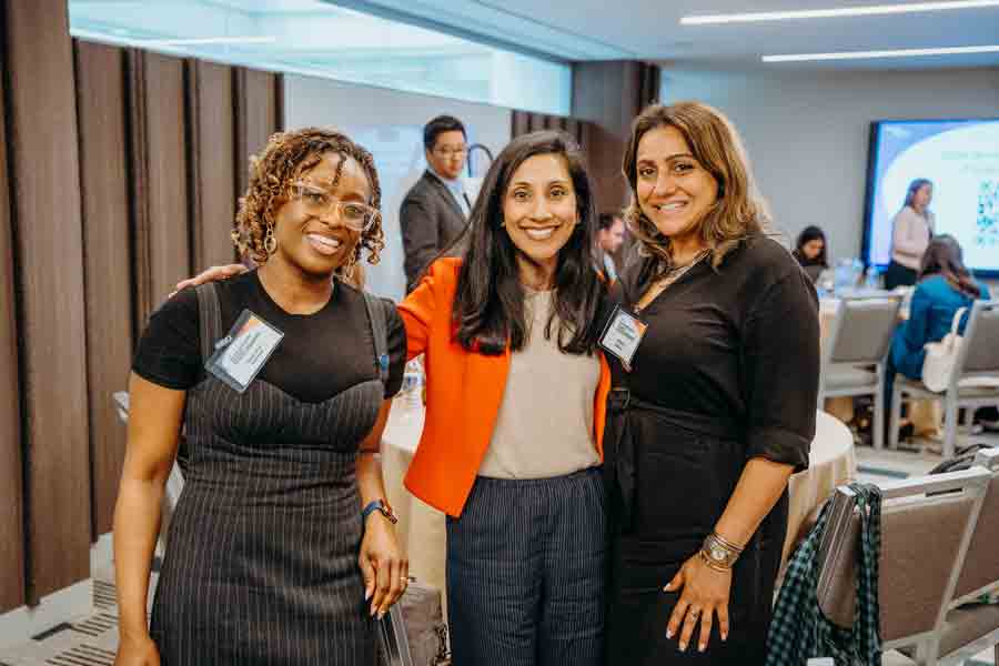 Three women stand together smiling at a conference or meeting room. Two women wear black outfits, while the center woman wears a red blazer. People are seated and talking at tables in the background.