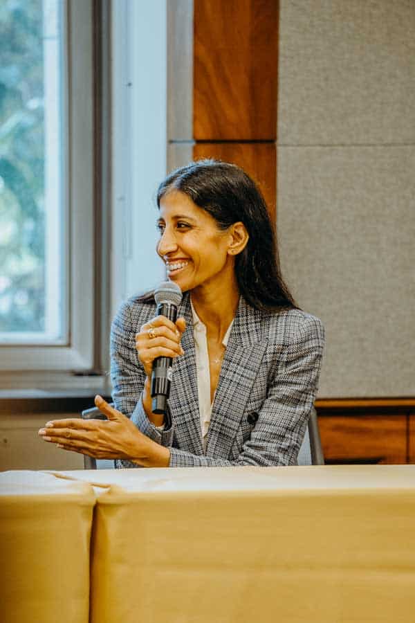 A woman in a plaid blazer sits at a table, smiling and holding a microphone while gesturing with her other hand. She appears to be speaking at an indoor event or panel discussion.